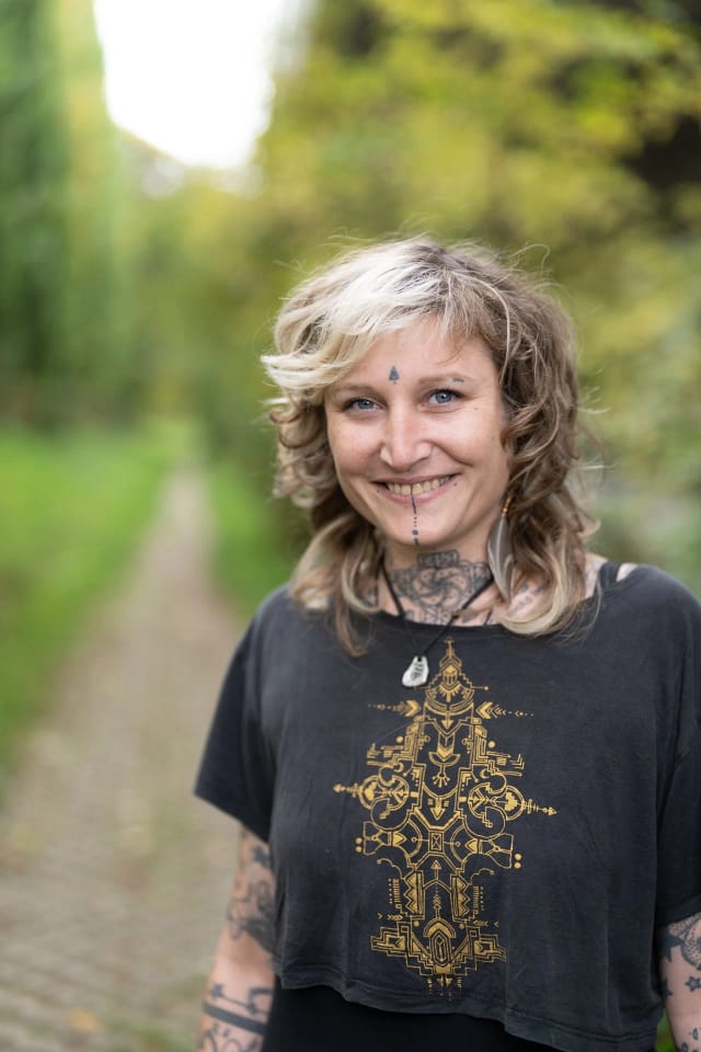 Smiling woman outdoors with tattoos, black T-shirt with gold pattern, multi-coloured short hair, green forest path in the background.